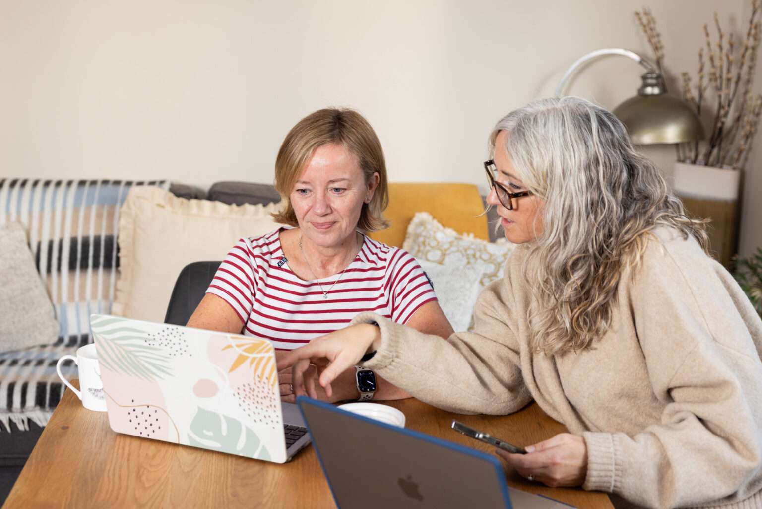 Two women sit at a wooden table with open laptops. One woman, in a striped shirt, looks at her laptop whilst the other, in glasses and a beige jumper, points at the screen. They appear to be discussing something.
