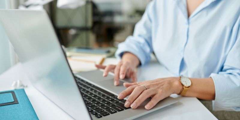 A person wearing a light blue shirt types on a laptop at a desk. A diary, pen, and some papers are visible nearby. The scene appears to be an office or home workspace.