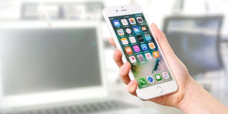 A hand holding a mobile phone with various app icons displayed on the screen. A blurred laptop and office environment are visible in the background.