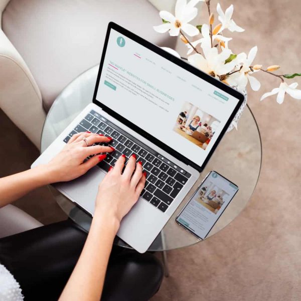 A person types on a laptop at a glass table with a mobile phone next to them. A flower arrangement and an armchair are visible nearby. The laptop screen displays a website.